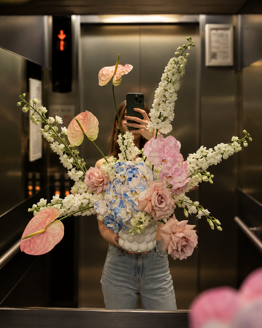 a large, vibrant floral arrangement with pink anthuriums, white delphiniums, pink roses, and blue hydrangeas in a textured white vase.