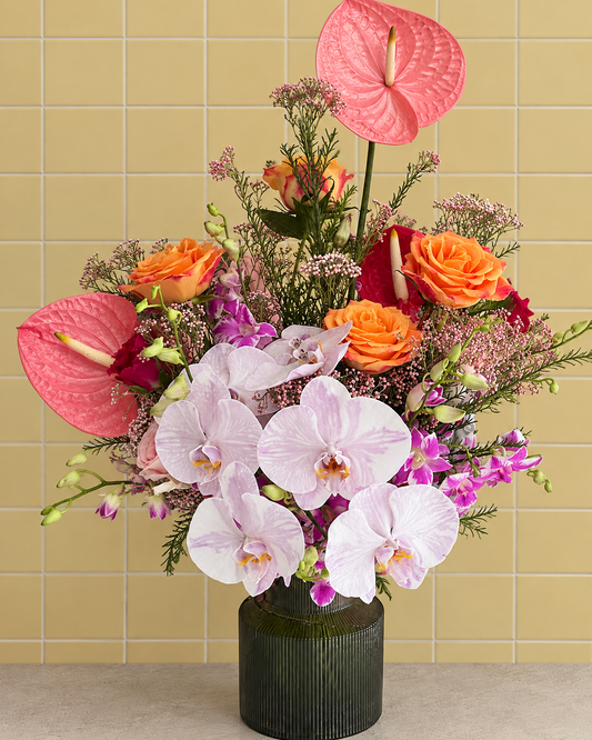 Deepavali flowers Singapore: Saffron arrangement of coral anthurium, orange roses and phalaenopsis orchids in a green glass vase