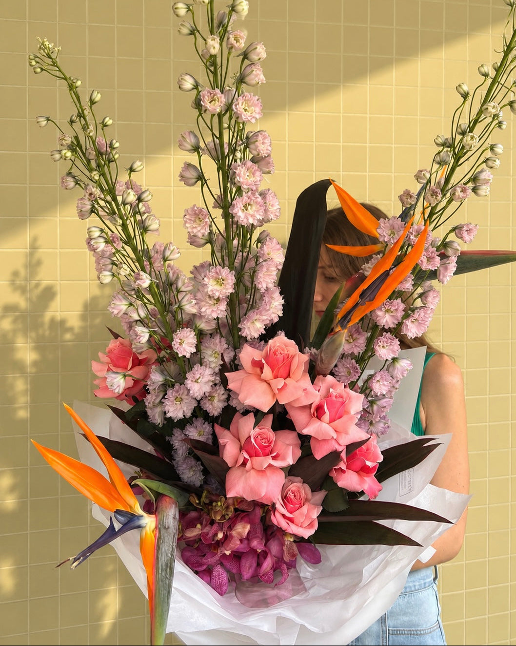 A large, vibrant floral arrangement held by a person against a beige tiled wall. The bouquet features light pink daisy-like flowers, bright pink roses, purple orchids, and orange bird of paradise flowers wrapped in white tissue paper.