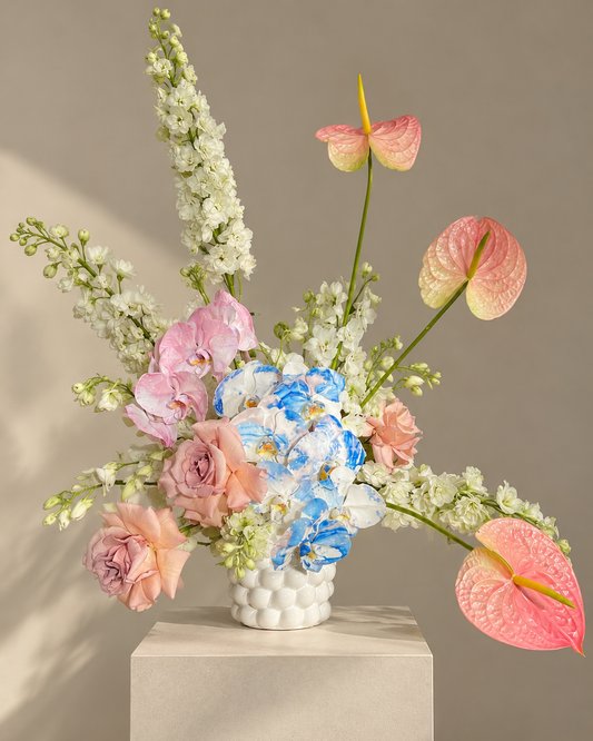 A vibrant floral arrangement featuring pink and peach roses, blue and white GLITCH orchids, pink anthuriums, and tall white delphiniums in a textured white ceramic vase by Gathered+Styled. Free delivery in Singapore.